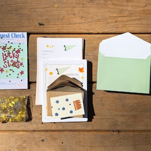 Op de afbeelding: Een assortiment briefpapier, waaronder een gastenbon met de tekst "Lucky Stars", een doorzichtige zak met gouden glitter en een lichtgroene envelop. Andere items zijn kaarten met illustraties en een bruine envelop, alles op een houten oppervlak.