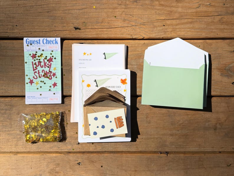 Op de afbeelding: Een assortiment briefpapier, waaronder een gastenbon met de tekst "Lucky Stars", een doorzichtige zak met gouden glitter en een lichtgroene envelop. Andere items zijn kaarten met illustraties en een bruine envelop, alles op een houten oppervlak.