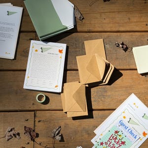 Op de afbeelding: Een bovenaanzicht van briefpapier op een houten oppervlak. Bevat een stapel groene en witte enveloppen, bruine enveloppen, een woordzoeker en een gastenbon met de tekst "Lucky Stars". Een rol washi-tape en een bloemensticker zijn ook zichtbaar.