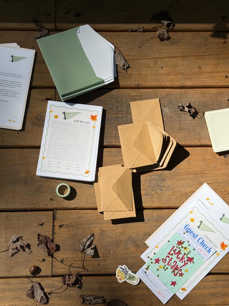 Op de afbeelding: Een bovenaanzicht van briefpapier op een houten oppervlak. Bevat een stapel groene en witte enveloppen, bruine enveloppen, een woordzoeker en een gastenbon met de tekst "Lucky Stars". Een rol washi-tape en een bloemensticker zijn ook zichtbaar.