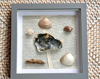 May include: A square gray shadow box frame containing a collage of vintage book pages layered with four assorted seashells and a small piece of driftwood. The shells vary in size, shape, and color, including tan, brown, and dark gray tones.