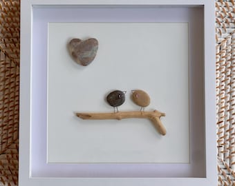 May include: A square white frame containing pebble art on a white background. The artwork features two small birds made of dark and light brown stones perched on a piece of driftwood, with a heart-shaped stone positioned above them.