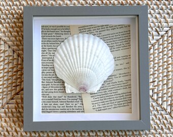 May include: A square gray frame containing a white scallop shell mounted over a background of vintage book pages with printed black text. The shell features a fan-shaped design with a subtle purple tint at the base, centered within the frame.
