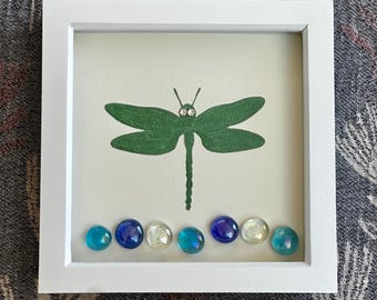 May include: A square white frame containing a cream-colored background with a centered green dragonfly illustration. The dragonfly features two small clear rhinestones as eyes. Below the dragonfly are eight smooth, round glass pebbles in blue.