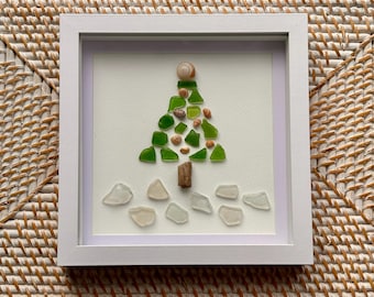 May include: A square white frame containing a piece of wall art featuring a tree shape made from green sea glass and small seashells. A piece of driftwood forms the trunk, and clear sea glass pieces are arranged at the base of the tree.