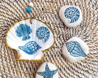 May include: A white scallop shell with a gold-painted rim and three blue shell prints, hanging from a jute twine with one wooden and two small blue beads, accompanied by three smooth white stones featuring blue nautical prints of shells and a starfish.