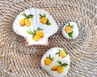 May include: A white scallop shell dish and two smooth white stones, all decorated with hand-painted yellow lemons and green leaves. The shell features a fluted edge and a cluster of lemons, while the stones display individual lemon motifs.