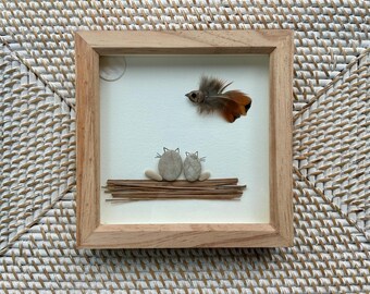 May include: A framed artwork featuring two pebble cats sitting on a bed of twigs, with a feathered bird and a circular moon. The artwork is set against a white background within a natural wood frame, creating a rustic aesthetic.