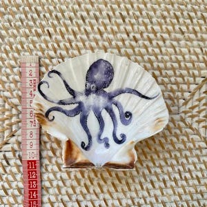 以下が含まれることがあります： 手描きの紫色のタコの模様が施された白いホタテ貝。貝殻の幅は約7.6cmで、自然で質感のある外観です。タコには詳細な触手と中央の頭があります。