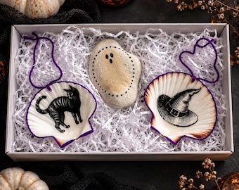 May include: A white gift box containing two scallop shell ornaments with purple string loops and a painted stone. One shell features a black cat illustration, the other a witch hat. The stone is painted with a ghost figure and black dotted outlines.
