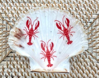 May include: A white scallop shell with three red lobster illustrations. The shell has brown and tan accents and sits on a woven, light brown surface. The lobsters are facing different directions.