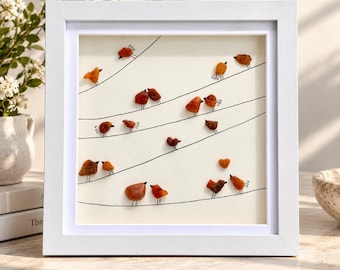 May include: A square white frame containing a piece of wall art on a cream background. The artwork features multiple small, orange-brown amber-like stones shaped as birds perched on thin black wire lines drawn across the surface.