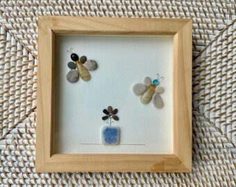 May include: A framed artwork featuring pebble art. Two pebble bees and a pebble flower in a blue square vase are arranged on a white background. The frame is made of light-colored wood.