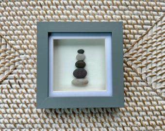 May include: A framed artwork featuring a stack of smooth, rounded stones in shades of gray and black. The stones are arranged vertically within a white and light blue mat, encased in a square gray frame. The artwork is displayed on a woven, textured surface.