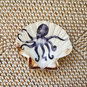 May include: A hand-painted octopus design on a white scallop shell. The octopus is depicted in shades of blue and purple, with detailed tentacles. The shell has a brown edge and is displayed on a woven surface.