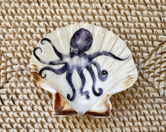 May include: A white scallop shell with a hand-painted octopus design in shades of blue and purple. The octopus has detailed tentacles and is centered on the shell. The shell has a brown edge and is displayed on a woven surface.