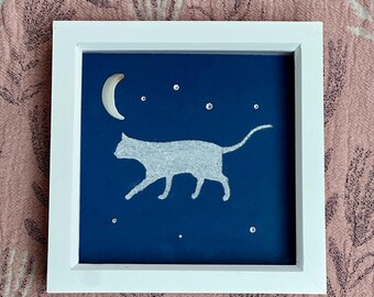 May include: A square white frame containing a dark blue background with a white silhouette of a walking cat. A crescent moon shape is positioned above the cat, and several small, clear, circular embellishments are scattered like stars.
