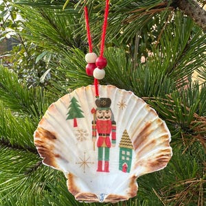 May include: A seashell Christmas decoration with a red string and wooden beads. The shell features a nutcracker, a Christmas tree, a house, and snowflakes. The shell has brown edges and is hanging from a pine tree branch.