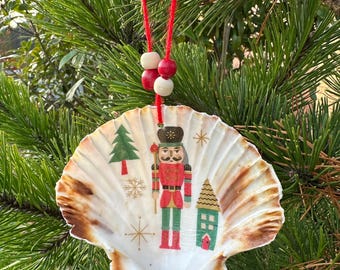 May include: A seashell Christmas ornament with a red string and wooden beads. The shell is decorated with a nutcracker, a Christmas tree, a house, and snowflakes. The shell has brown edges and is hanging from a pine tree branch.