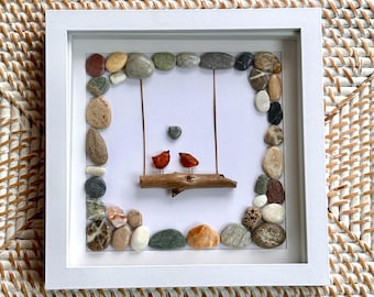 May include: A square white frame containing pebble art featuring two small orange birds perched on a wooden branch swing suspended by thin wires. A small grey heart-shaped stone is positioned above the birds. The border is lined with various smooth, multi-colored natural stones.