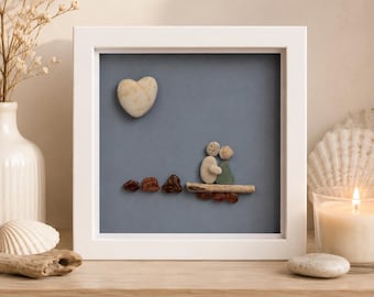 May include: A square white frame containing pebble art on a blue background. The artwork features two figures made of small stones sitting on a piece of driftwood, with a heart-shaped stone above them and several dark brown stones arranged below.