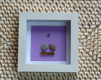 May include: A framed artwork featuring two pebble birds perched on a small branch against a purple background. A heart-shaped gem is above the birds. The artwork is encased in a white frame, perfect for home decor.