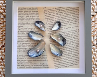 May include: A square gray frame containing a collage of vintage book pages with printed text. Five dark blue mussel shells are arranged in a circular pattern on the paper, each featuring a small, round, light pink pearl centered inside the shell.