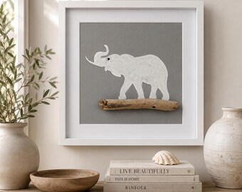 May include: A square white frame containing a gray background with a white elephant silhouette standing on a piece of driftwood. The elephant has a small black eye detail. The artwork is presented as a wall-mounted piece of decor.