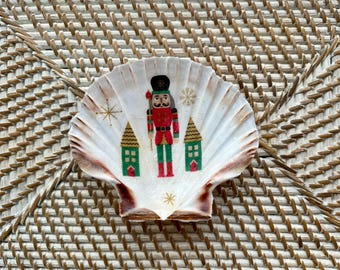 May include: A decorative seashell with a Christmas theme. The shell features a painted nutcracker soldier, two green houses, and gold star accents. The shell has a natural white and brown color.