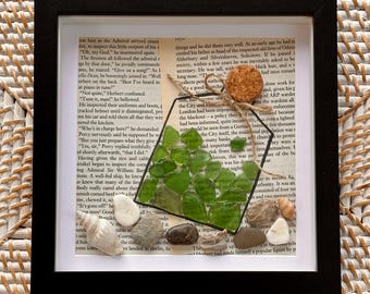 May include: A square black frame containing a collage on a vintage book page. The artwork features a bottle outline filled with green sea glass, topped with a cork and twine. Several small seashells and smooth pebbles are arranged at the base.