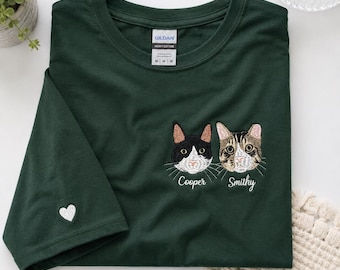 May include: A dark green short-sleeve t-shirt featuring embroidered portraits of two cats on the chest, labeled Cooper and Smithy. A small white heart is embroidered on the sleeve. The shirt has a Gildan brand tag at the neck showing size M.