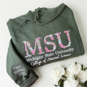 Puede incluir: Una sudadera con capucha verde oliva con las letras "MSU" bordadas en un diseño floral. Debajo de las letras están las palabras "Michigan State University" y "College of Natural Science". La manga dice "Plant Biology 2025".