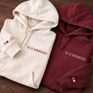 May include: Two hooded sweatshirts, one cream-colored and one burgundy. The cream-colored sweatshirt has the initials "P" and a heart on the cuff, and the Roman numeral date "IV.X.MMXXVI". The burgundy sweatshirt has the initials "K" and a heart on the cuff, and the Roman numeral date "IV.X.MMXXVI".