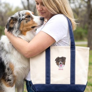 A woman hugging her dog while wearing a personalized Natural/Navy boat tote featuring a custom pet embroidery portrait. This sentimental dog mom tote bag showcases the large capacity and durable straps of the Liberty Bags 8871, making it a great customizable gift.