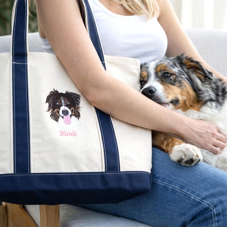 A woman relaxing on a sofa with her dog and a Natural/Navy Bags 8871 personalized tote. The custom embroidered dog face adds a unique touch to this classic canvas boat tote. Search-optimized for personalized pet keepsakes and custom embroidery fashion.