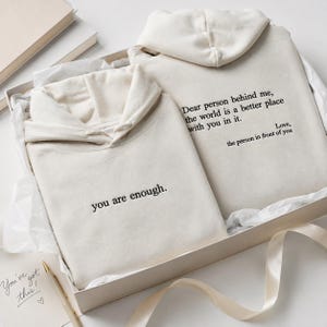 Op de afbeelding: Twee crèmekleurige hoodies opgevouwen in een geschenkdoos. Eén hoodie is voorzien van de zwarte geborduurde tekst 'you are enough.' De andere hoodie toont de zwarte geborduurde tekst 'Dear person behind me, the world is a better place with you in it. Love, the person in front of you.'