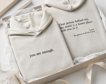 Könnte beinhalten: Zwei cremefarbene Kapuzenpullover, die in einer Geschenkbox gefaltet sind. Ein Hoodie zeigt den schwarzen gestickten Text 'you are enough.' Der andere Hoodie zeigt den schwarzen gestickten Text 'Dear person behind me, the world is a better place with you in it. Love, the person in front of you.'