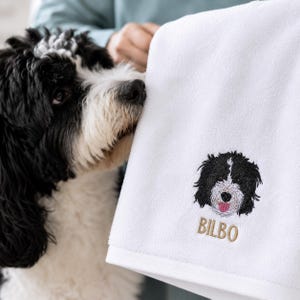May include: White dog towel with an embroidered black and white dog face and the name "BILBO" in gold thread. The towel is soft and fluffy, perfect for pet owners.
