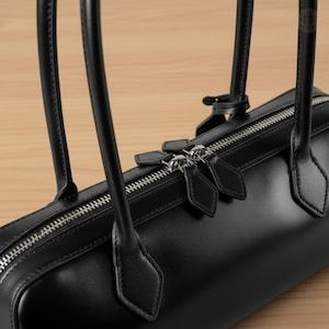 May include: A black leather handbag with a cylindrical shape, featuring a silver zipper and dual handles. The bag has a minimalist design, with a focus on quality materials and craftsmanship. The handles are attached with diamond-shaped leather accents.