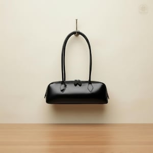 May include: A black leather handbag with a rectangular shape and two long, curved handles. The bag is suspended from a metal hook against a cream-colored wall. The bag has a zipper closure and diamond-shaped accents.