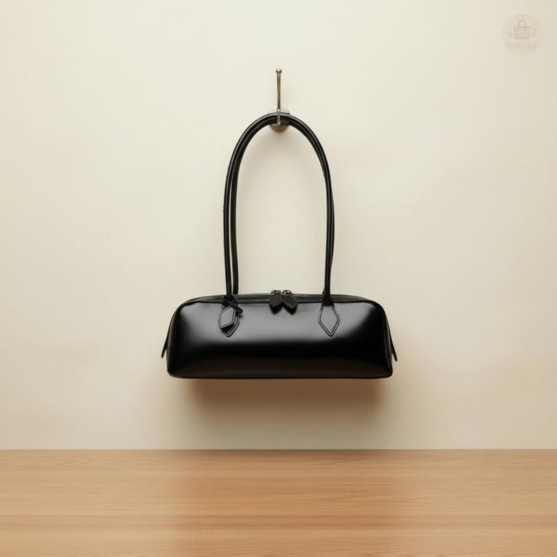 May include: A black leather handbag with a rectangular shape and two long, curved handles. The bag is suspended from a metal hook against a cream-colored wall. The bag has a zipper closure and diamond-shaped accents.