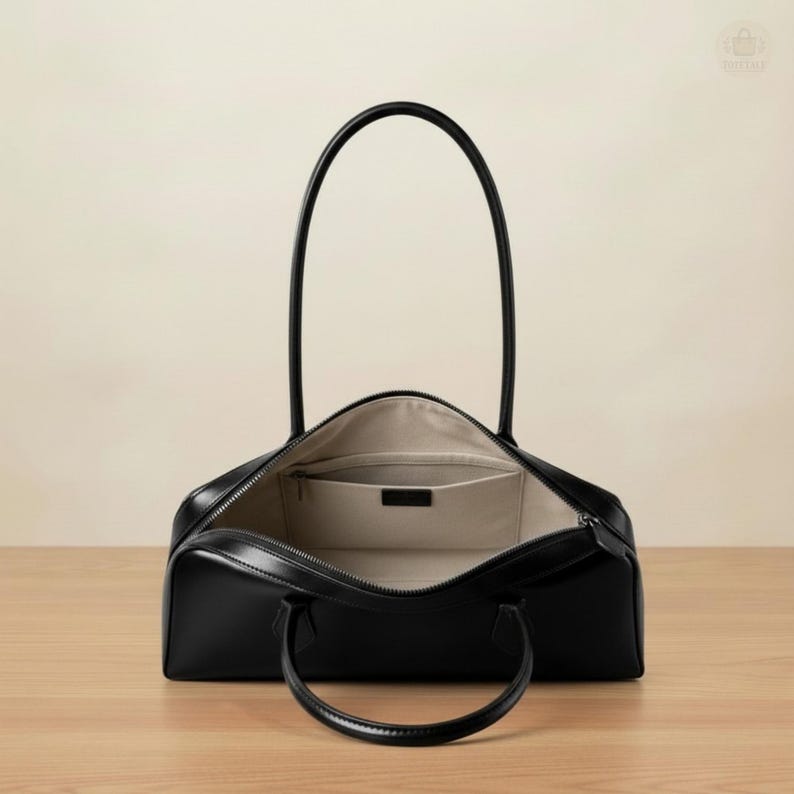 May include: Black leather tote bag with a rectangular shape and two rounded handles. The bag is open, revealing a light-colored interior with a small pocket. The zipper is open, and the bag sits on a wooden surface.