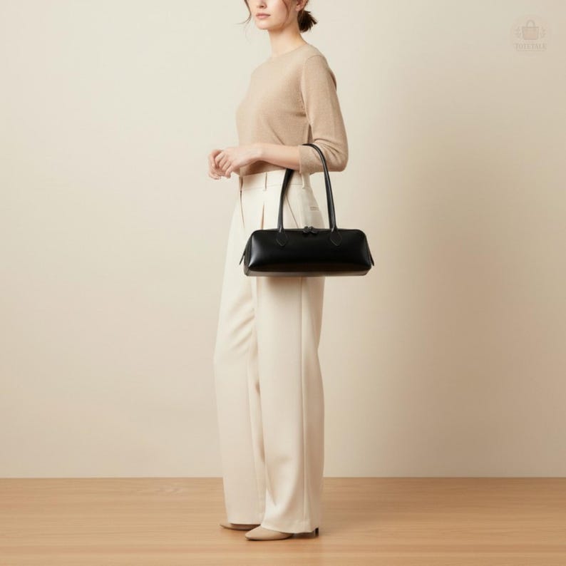 May include: A black rectangular handbag with two handles. The bag is held by a person wearing a beige sweater and white pants. The bag has a zipper closure and a sleek, minimalist design.