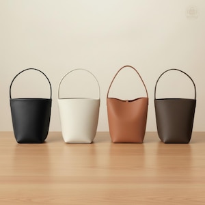 May include: Four leather bucket bags in black, white, tan, and dark brown, each with a matching handle. The bags are displayed on a light wood surface against a neutral background. The bags have a simple, minimalist design.