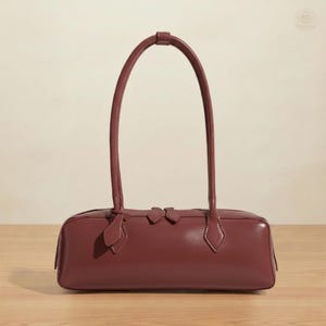 May include: A burgundy-colored rectangular handbag with two long, arched handles. The bag features a zipper closure and decorative accents. The bag is made of a smooth material and sits on a wooden surface.