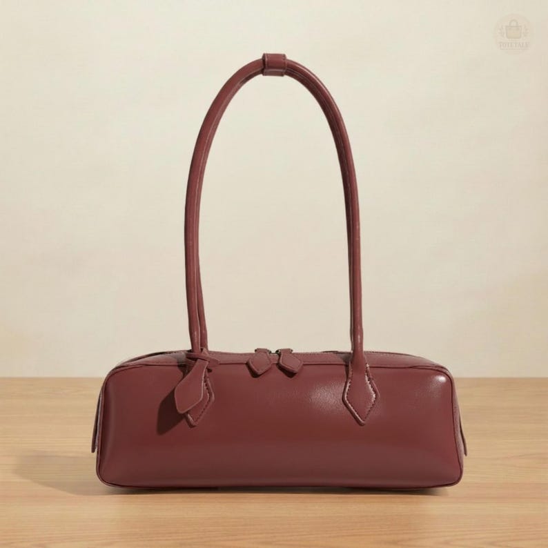 May include: A burgundy-colored rectangular handbag with two long, arched handles. The bag features a zipper closure and decorative accents. The bag is made of a smooth material and sits on a wooden surface.