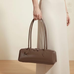 May include: A brown rectangular handbag with long, thin handles. The bag has a structured shape with a subtle design on the front. The handles are attached to the bag with decorative elements. The bag is held by a person wearing a cream-colored dress.