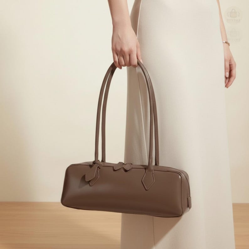 May include: A brown rectangular handbag with long, thin handles. The bag has a structured shape with a subtle design on the front. The handles are attached to the bag with decorative elements. The bag is held by a person wearing a cream-colored dress.