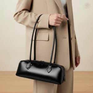 May include: A sleek, black leather handbag with a rectangular shape and long, slender handles. The bag features a zipper closure and small decorative accents. The glossy finish of the bag contrasts with the neutral-toned clothing in the background.