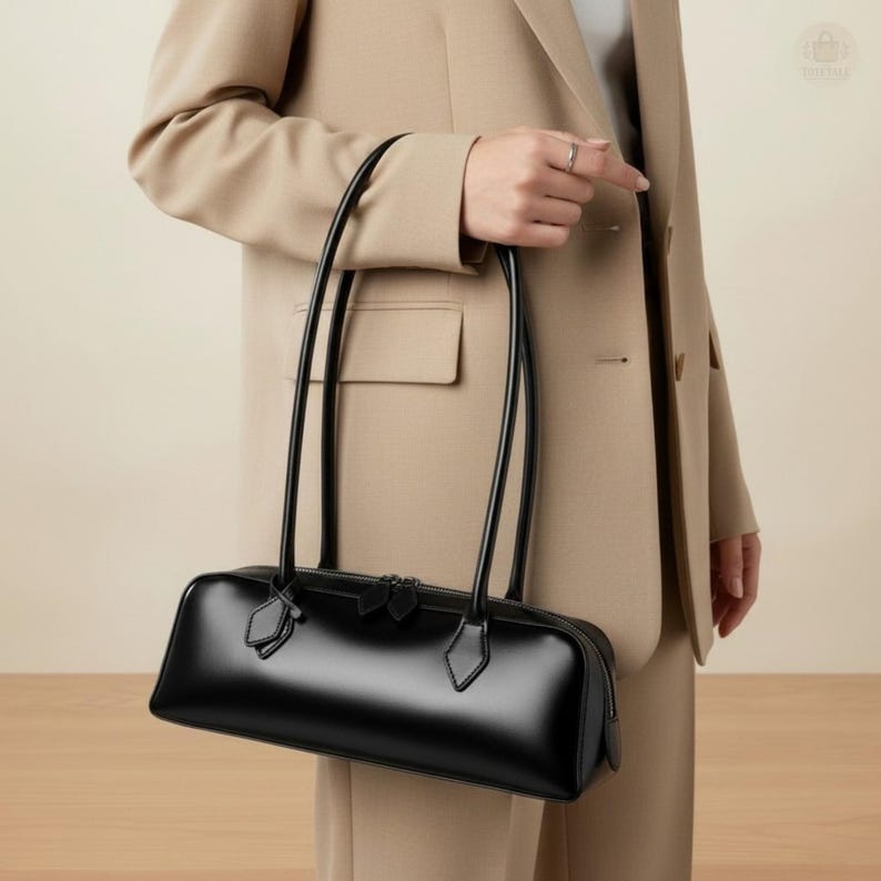 May include: A sleek, black leather handbag with a rectangular shape and long, slender handles. The bag features a zipper closure and small decorative accents. The glossy finish of the bag contrasts with the neutral-toned clothing in the background.